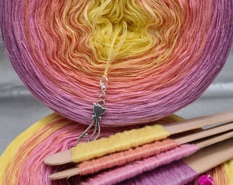May include: A skein of yellow, orange, and pink ombre yarn with a silver charm attached. The yarn is wound around a cardboard tube and there are three wooden knitting needles with yarn wrapped around them.