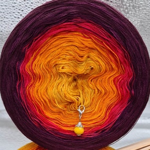 May include: A ball of yarn with a gradient of purple, red, orange, and yellow. A silver stitch marker with a yellow bead is attached to the yarn.