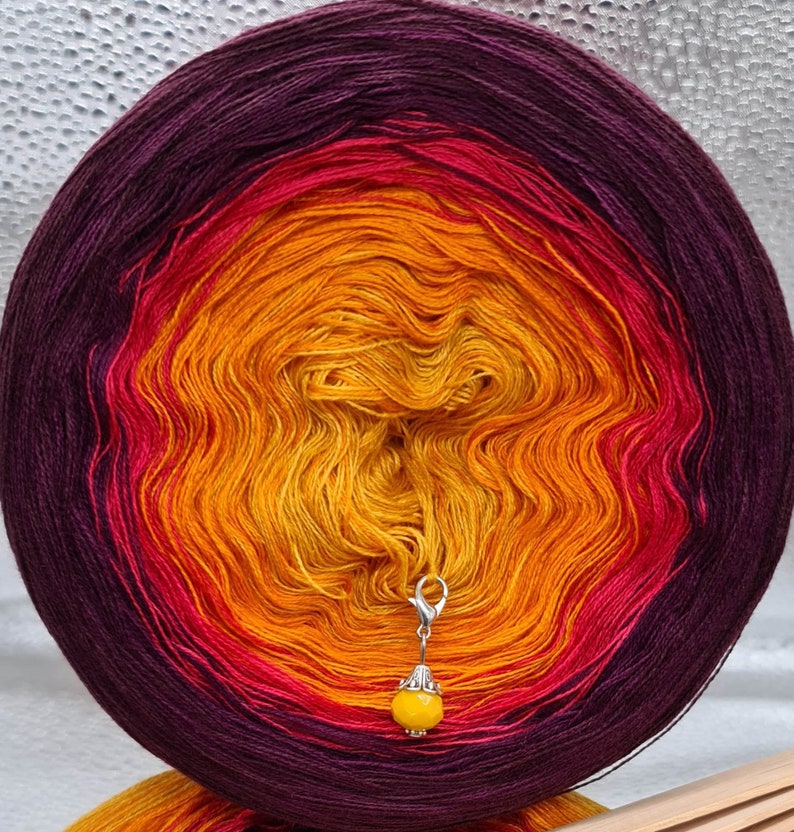 May include: A ball of yarn with a gradient of purple, red, orange, and yellow. A silver stitch marker with a yellow bead is attached to the yarn.