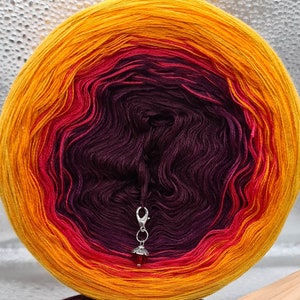 May include: A skein of yarn in a gradient of orange, red, and purple colors. The yarn is wound into a ball shape and has a silver yarn needle attached to it.
