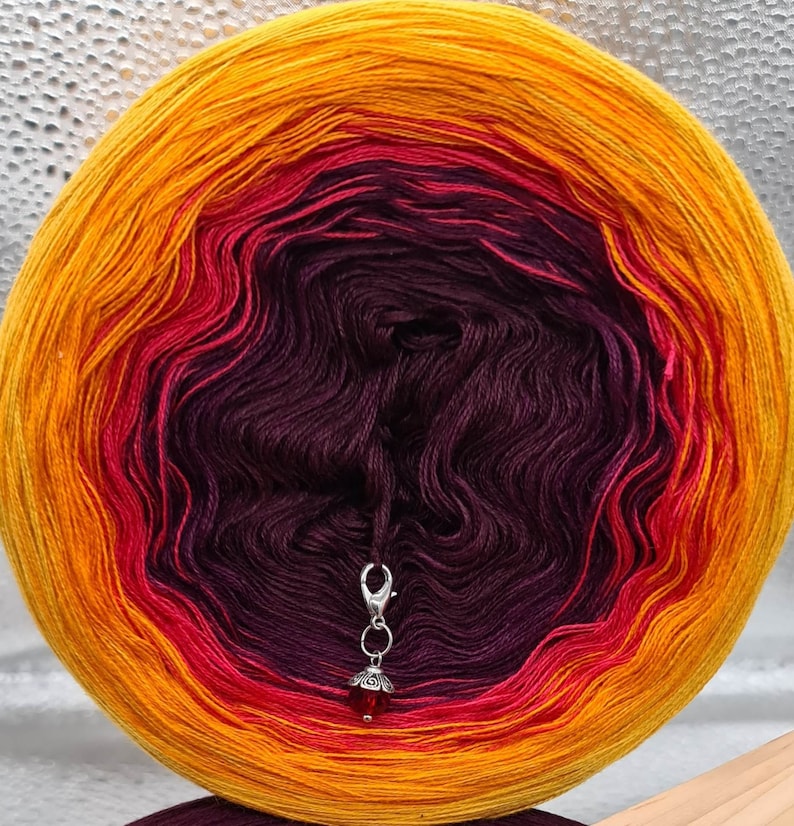 May include: A skein of yarn in a gradient of orange, red, and purple colors. The yarn is wound into a ball shape and has a silver yarn needle attached to it.