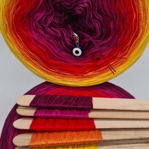 May include: A skein of yarn in a gradient of purple, red, orange, and yellow colours. The yarn is wound around a white cardboard tube. The yarn is displayed on a wooden dowel with the different colours of the yarn separated.