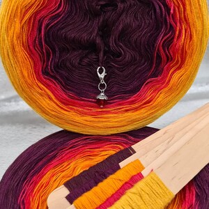 May include: Two skeins of yarn in a gradient of purple, red, and orange. The yarn is wound into balls and the skeins are on a white surface. The yarn is wrapped around wooden sticks.