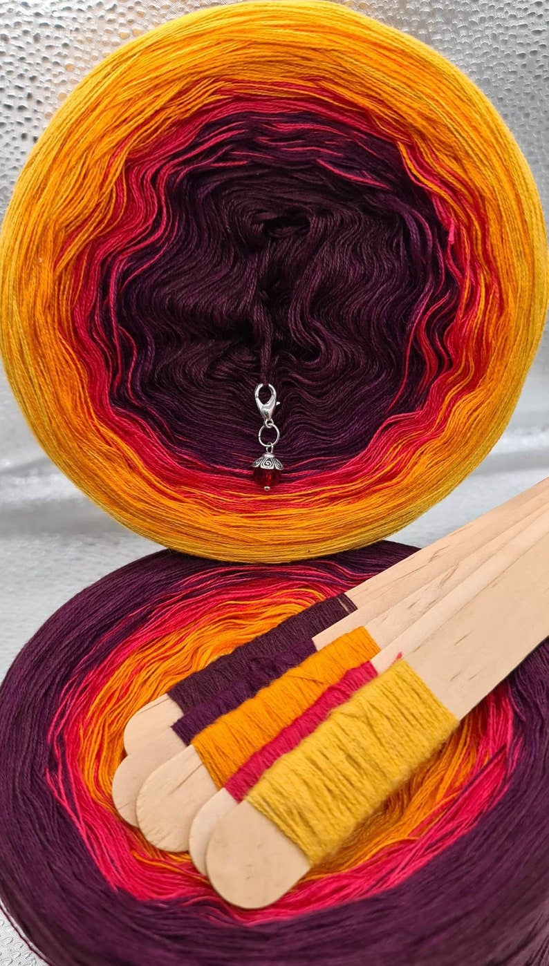 May include: Two skeins of yarn in a gradient of purple, red, and orange. The yarn is wound into balls and the skeins are on a white surface. The yarn is wrapped around wooden sticks.