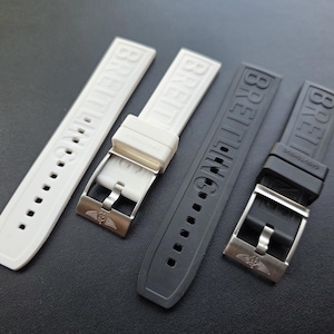 Könnte beinhalten: Vier Uhrenarmbänder in Weiß und Schwarz. Die Armbänder sind aus Gummi und tragen den Markennamen "BREITLING" geprägt. Jedes Armband hat eine silberfarbene Schnalle mit einem Logo.