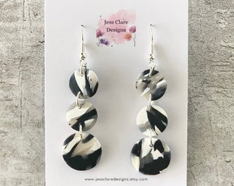 May include: Black and white patterned dangle earrings with three round discs on each earring. The earrings are silver and have a hook closure. The earrings are displayed on a white card with the text "Jess Clare Designs" and a website address.