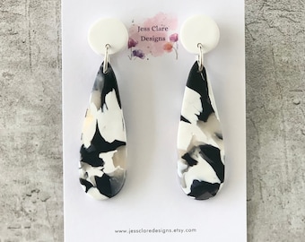 May include: A pair of black and white teardrop-shaped earrings with a white circle at the top. The earrings are made of polymer clay and have a marbled effect. The earrings are on a white card with the text "Jess Clare Designs" and a website address.