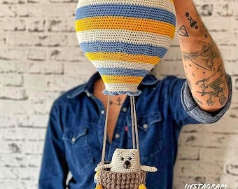 Può includere: Un pallone aerostatico all'uncinetto con strisce gialle e blu, che tiene un animale all'uncinetto in un cestino. Il pallone è sospeso da delle corde ed è tenuto da una mano. L'immagine include il testo "Instagram herr.wolle".