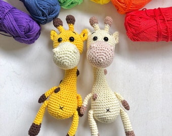 May include: Two crocheted giraffe toys, one yellow and one cream-colored, with brown spots and accents. The giraffes are surrounded by colorful skeins of yarn in a rainbow arrangement, including purple, blue, yellow, orange, and red.