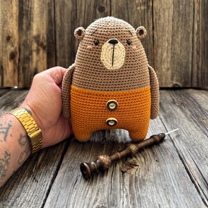 May include: A crocheted brown bear wearing orange pants with two white buttons. The bear is sitting on a wooden surface.