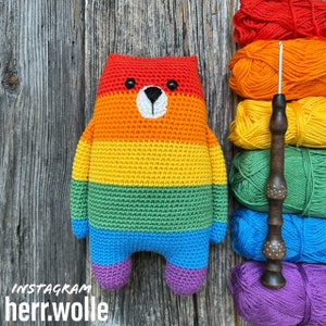 May include: A crocheted rainbow bear with a white face and black eyes. The bear is sitting on a wooden surface with balls of yarn in the background.