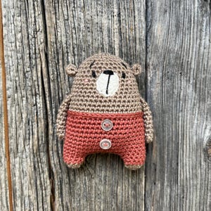 May include: A crocheted brown bear wearing red pants with two white buttons. The bear is standing against a wooden background.