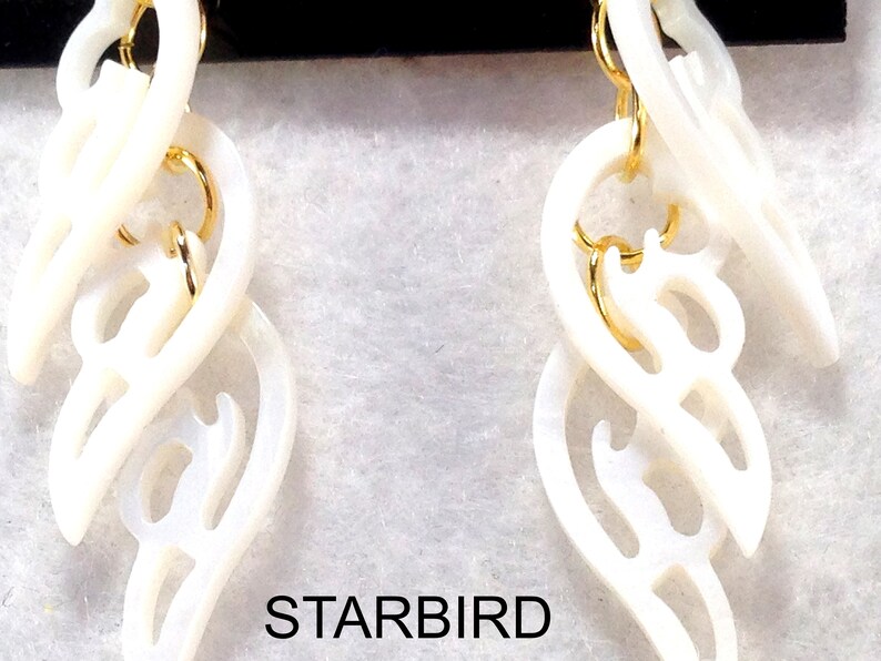 Puede incluir: Un par de pendientes de concha blanca con una cadena dorada y un dise&ntilde;o delicado e intrincado. Los pendientes tienen forma de una serie de corazones superpuestos.