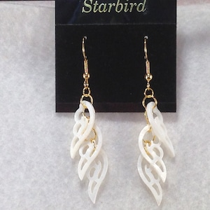 Puede incluir: Un par de pendientes de concha blanca con ganchos dorados y un dise&ntilde;o delicado e intrincado. Los pendientes se muestran en una tarjeta negra con la palabra "Starbird" impresa en ella.