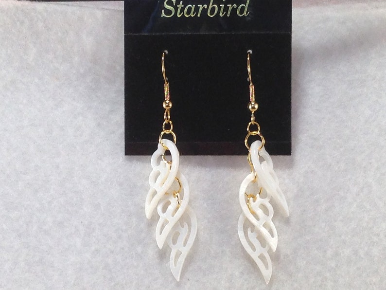 Puede incluir: Un par de pendientes de concha blanca con ganchos dorados y un dise&ntilde;o delicado e intrincado. Los pendientes se muestran en una tarjeta negra con la palabra "Starbird" impresa en ella.