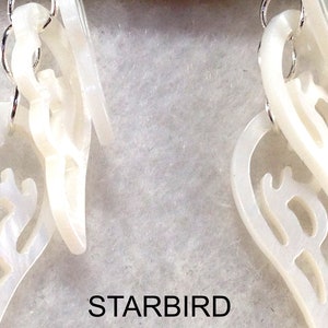 Puede incluir: Un par de pendientes blancos en forma de concha con intrincados recortes. Los pendientes est&aacute;n suspendidos de ganchos plateados.