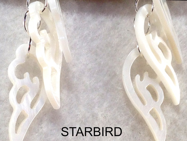 Puede incluir: Un par de pendientes blancos en forma de concha con intrincados recortes. Los pendientes est&aacute;n suspendidos de ganchos plateados.