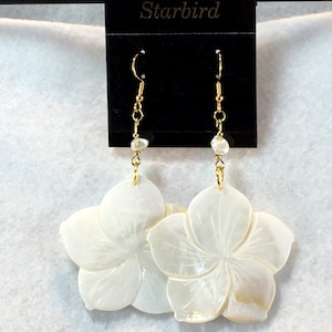 May include: A pair of white shell flower earrings with gold accents. The earrings are hanging from a black card with the word "Starbird" printed on it.