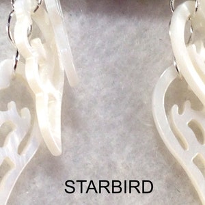 Puede incluir: Un par de pendientes blancos en forma de concha con intrincados recortes. Los pendientes cuelgan de ganchos plateados.