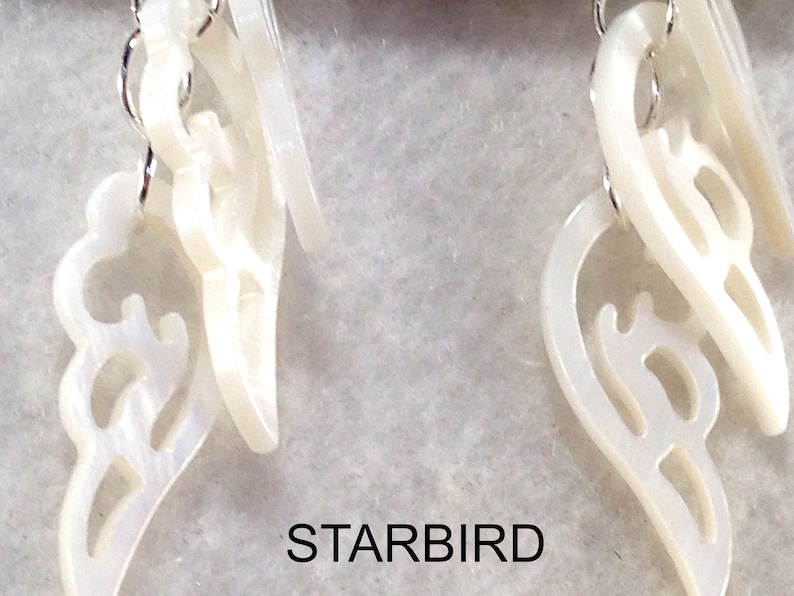 Puede incluir: Un par de pendientes blancos en forma de concha con intrincados recortes. Los pendientes cuelgan de ganchos plateados.