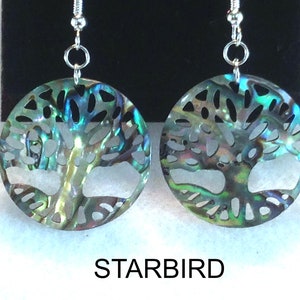 May include: A pair of iridescent abalone shell earrings. Each earring features a circular design with a tree of life cut-out. The earrings hang from silver-colored hooks. The word "STARBIRD" is printed below the earrings.