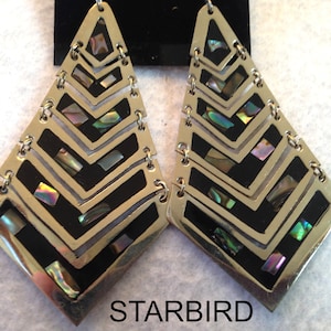 May include: A pair of gold-tone metal dangle earrings featuring a geometric chevron pattern with black enamel inlays and iridescent abalone shell accents. The earrings are displayed on a black card with the text STARBIRD printed in white.