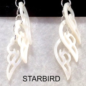 Puede incluir: Un par de pendientes blancos en forma de concha con intrincados recortes. Los pendientes est&aacute;n conectados por una cadena plateada.