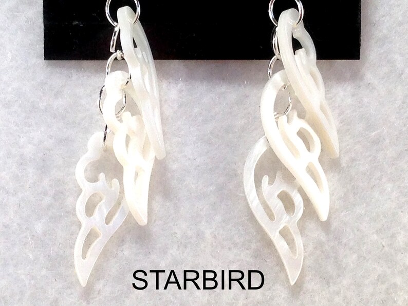 Puede incluir: Un par de pendientes blancos en forma de concha con intrincados recortes. Los pendientes est&aacute;n conectados por una cadena plateada.