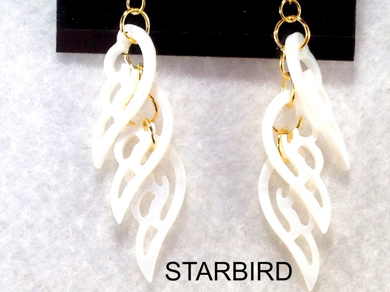 Puede incluir: Un par de pendientes blancos en forma de concha con detalles en metal dorado. Los pendientes est&aacute;n conectados por una cadena y tienen un dise&ntilde;o delicado e intrincado.