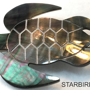 May include: A black and iridescent mother of pearl turtle brooch with a detailed shell design. The brooch is a decorative accessory for clothing.