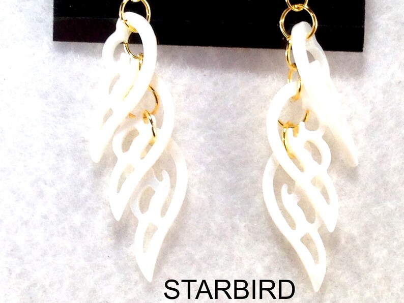 Puede incluir: Un par de pendientes blancos, tallados de forma intrincada, con detalles dorados. Los pendientes tienen forma de una serie de hojas superpuestas.