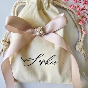 May include: A beige drawstring bag with a blush pink ribbon tied in a bow. The bow is adorned with a small, decorative brooch featuring pearls. The name "Sophie" is written in elegant script on the bag.