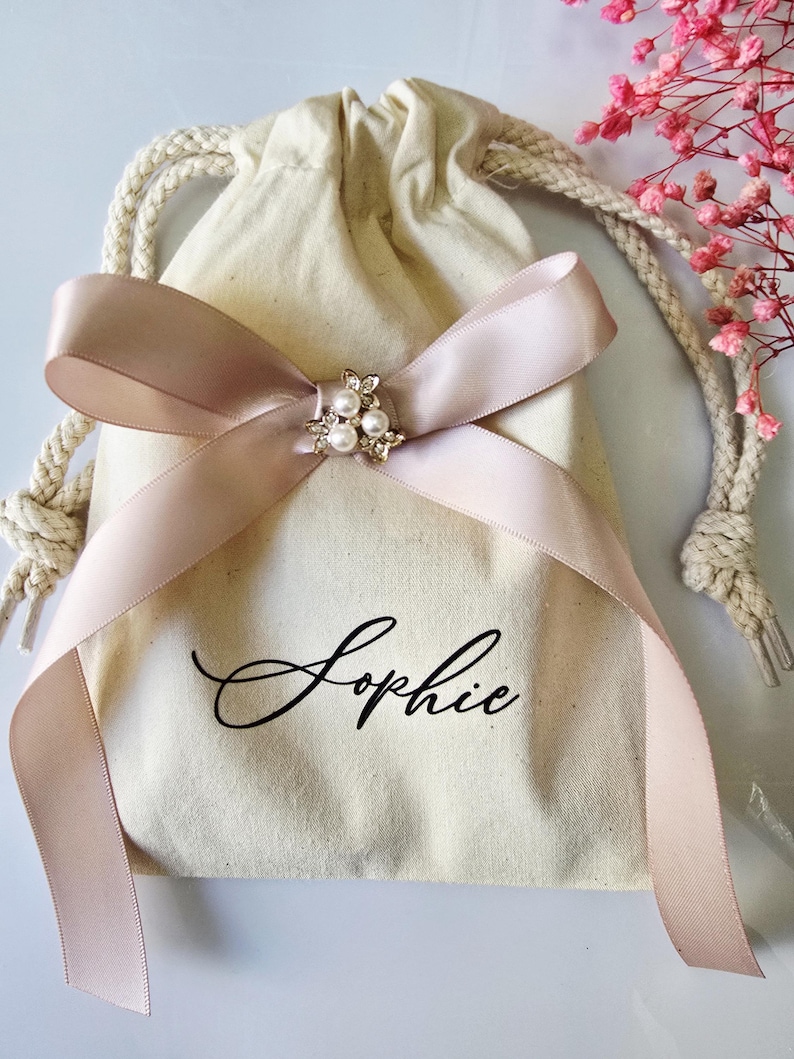 May include: A beige drawstring bag with a blush pink ribbon tied in a bow. The bow is adorned with a small, decorative brooch featuring pearls. The name "Sophie" is written in elegant script on the bag.