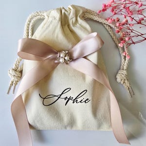 May include: A cream-colored drawstring bag with a blush pink satin ribbon tied in a bow. The bow is adorned with a silver-toned brooch with pearls. The name "Sophie" is written in elegant script on the bag.