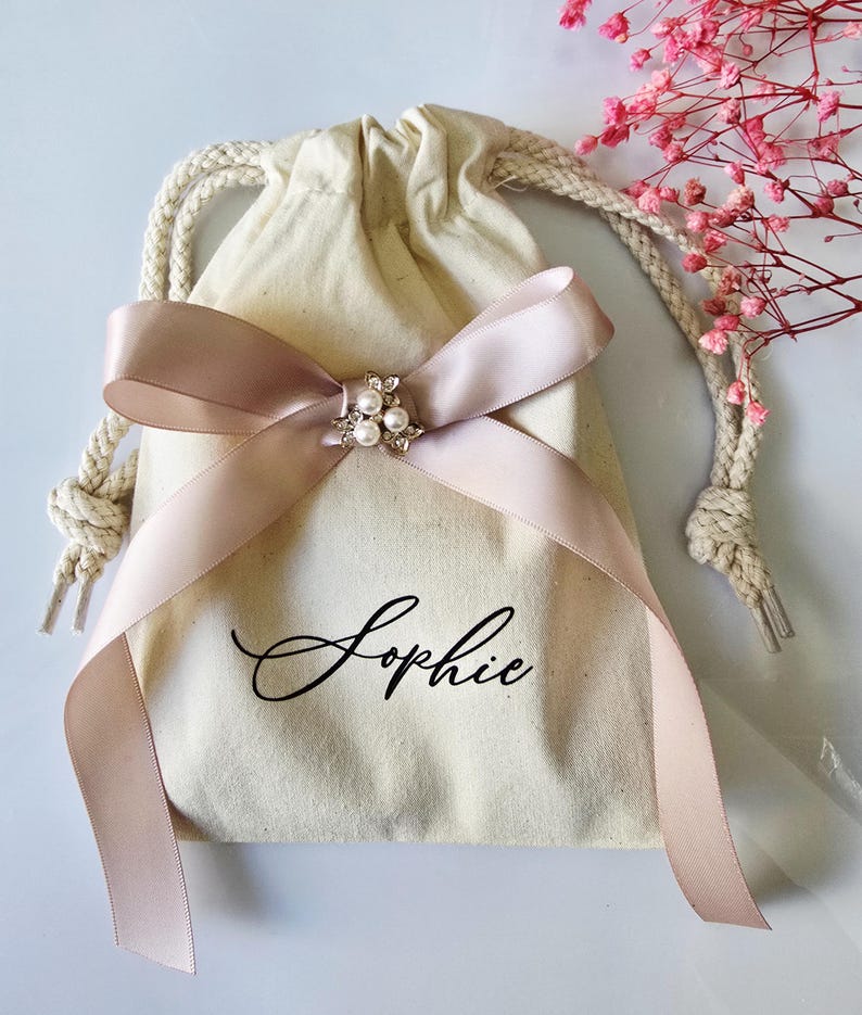 May include: A cream-colored drawstring bag with a blush pink satin ribbon tied in a bow. The bow is adorned with a silver-toned brooch with pearls. The name "Sophie" is written in elegant script on the bag.