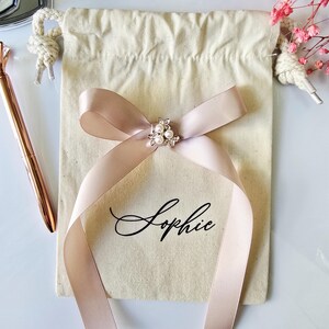 May include: A beige drawstring bag with a blush pink ribbon tied in a bow. The bow is adorned with a small, pearl-centered embellishment. The name "Sophie" is written in elegant script on the bag. A rose gold pen and pink flowers are in the background.
