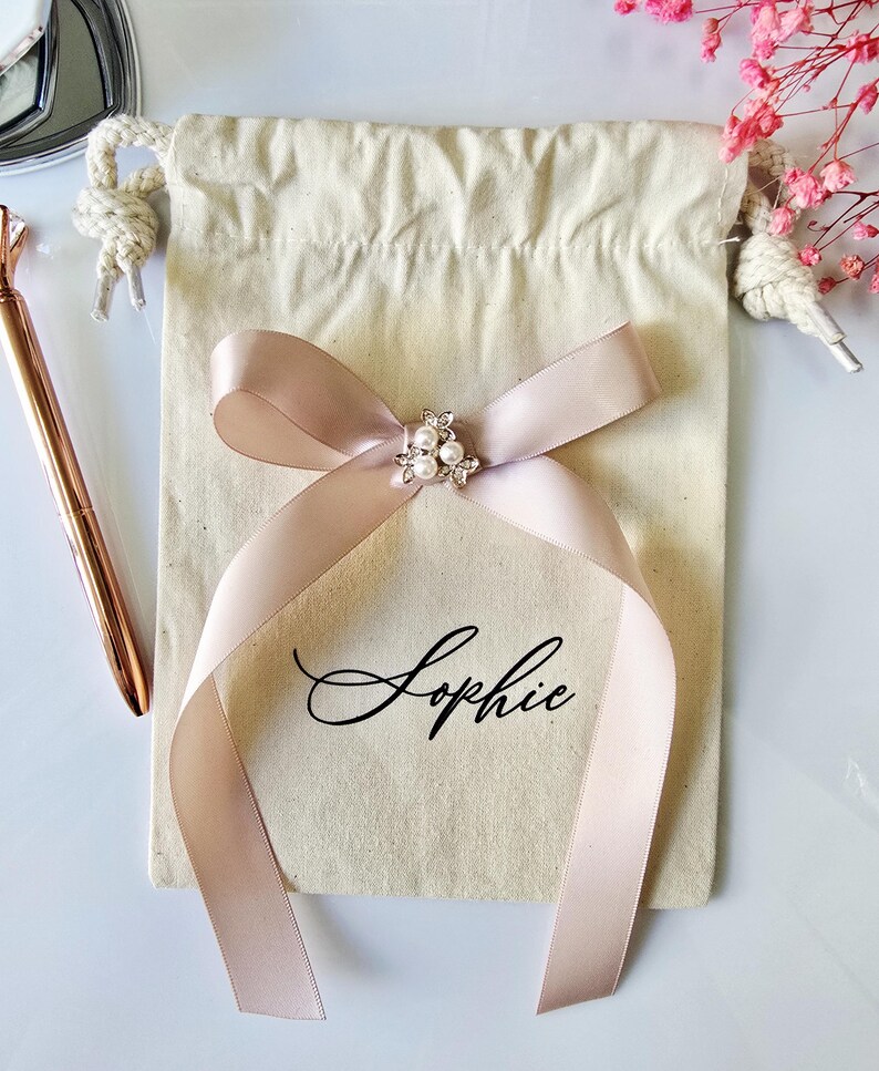 May include: A beige drawstring bag with a blush pink ribbon tied in a bow. The bow is adorned with a small, pearl-centered embellishment. The name "Sophie" is written in elegant script on the bag. A rose gold pen and pink flowers are in the background.