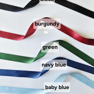 May include: Satin ribbons in black, burgundy, green, navy blue, and baby blue are displayed against a white background. Each ribbon is labeled with its color name. The ribbons are slightly curled, showcasing their texture and sheen.