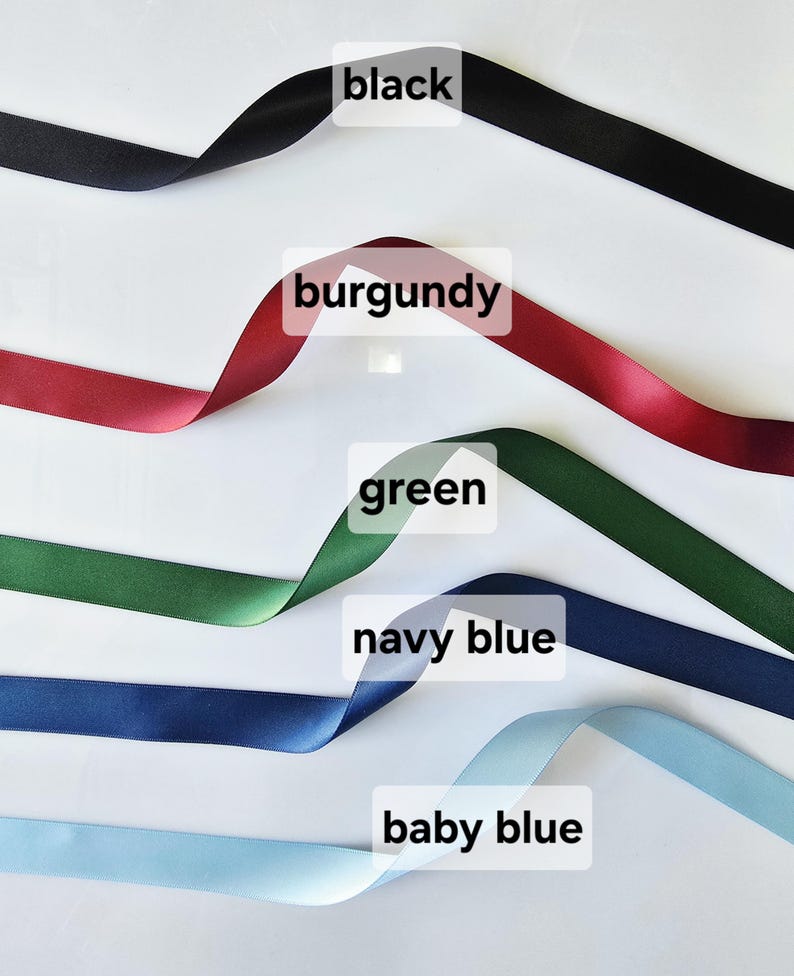 May include: Satin ribbons in black, burgundy, green, navy blue, and baby blue are displayed against a white background. Each ribbon is labeled with its color name. The ribbons are slightly curled, showcasing their texture and sheen.