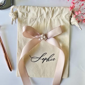 May include: A beige drawstring bag with a blush pink ribbon tied in a bow. The bow is adorned with a pearl and rhinestone embellishment. The name "Sophie" is printed in black script on the bag.
