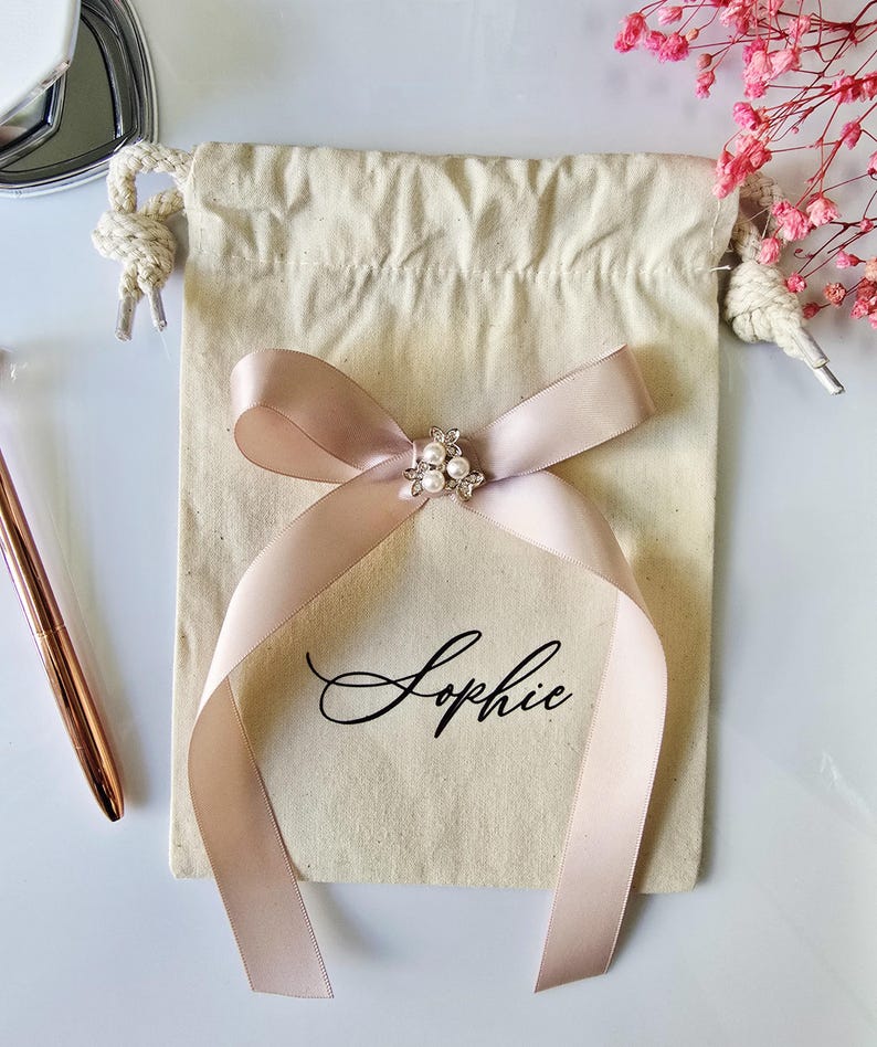 May include: A beige drawstring bag with a blush pink ribbon tied in a bow. The bow is adorned with a pearl and rhinestone embellishment. The name "Sophie" is printed in black script on the bag.