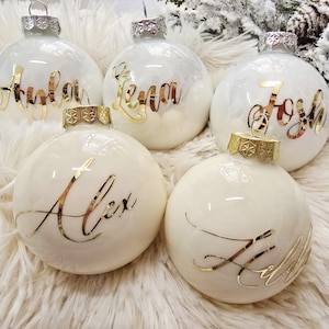 Könnte beinhalten: Fünf weiße Weihnachtskugeln mit goldenen Buchstaben, die Namen buchstabieren. Jede Kugel hat eine silberne oder goldene Kappe. Die Kugeln sind auf einer flauschigen weißen Oberfläche angeordnet und schaffen eine festliche Urlaubsszene.