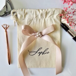 May include: A beige drawstring gift bag with a blush pink ribbon tied in a bow. The bow is adorned with a small, decorative cluster of pearls and crystals. The name "Sophie" is printed on the bag in elegant script. A rose gold pen and lipstick are also visible.