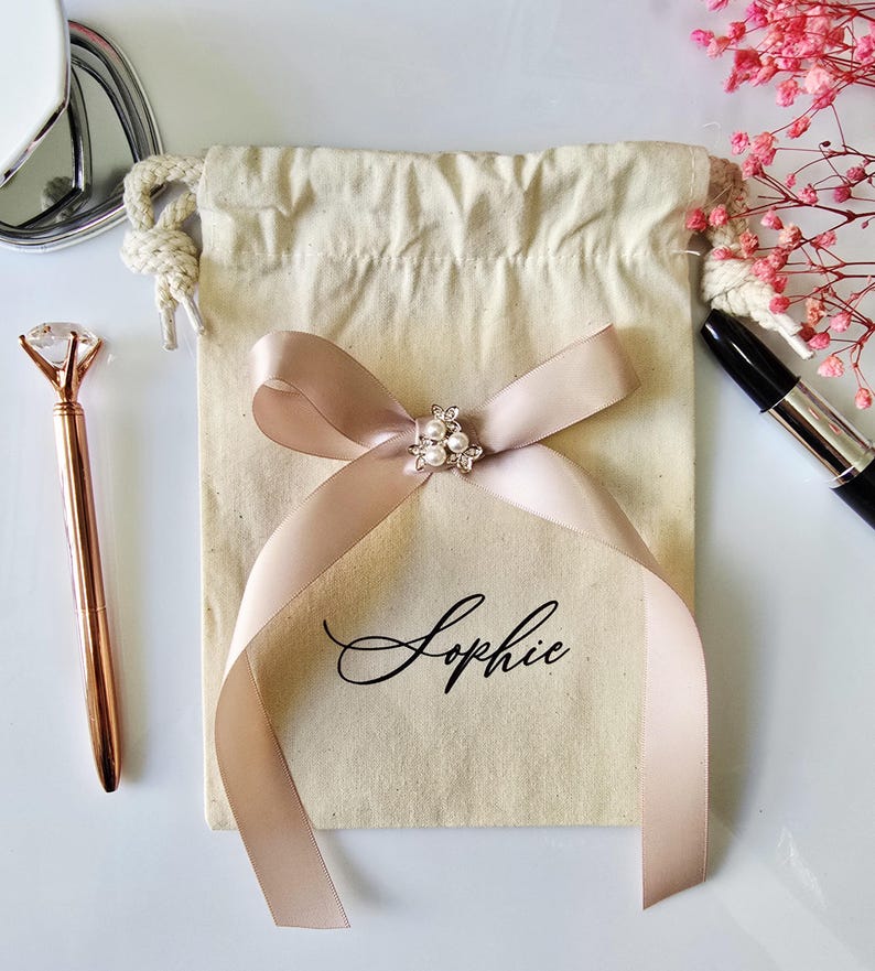 May include: A beige drawstring gift bag with a blush pink ribbon tied in a bow. The bow is adorned with a small, decorative cluster of pearls and crystals. The name "Sophie" is printed on the bag in elegant script. A rose gold pen and lipstick are also visible.