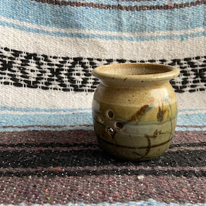 Puede incluir: Un pequeño jarrón de cerámica hecho a mano con un esmalte moteado de color beige. El jarrón tiene un cuerpo redondeado con tres pequeños agujeros y un borde ligeramente ensanchado. Diseños abstractos marrones y azules decoran la superficie. El jarrón está sobre un textil a rayas.