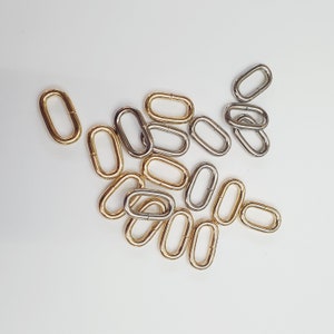 Peut inclure: Un groupe d'anneaux métalliques ovales en tons or et argent. Les anneaux sont disposés en un tas lâche sur une surface blanche.