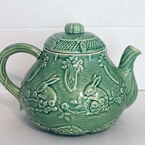 Peut inclure: Une théière en céramique vert sauge ornée d'un relief représentant des lapins, des carottes et des vignes feuillues. La théière possède un couvercle assorti avec un motif en treillis texturé, une anse et un bec verseur courbés, le tout vernissé.