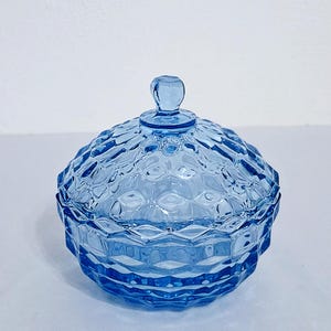 May include: A light blue glass candy dish with a matching lid, featuring a raised diamond-shaped geometric pattern across the exterior surface. The circular container has a small, rounded knob handle on top of the domed lid for easy removal.