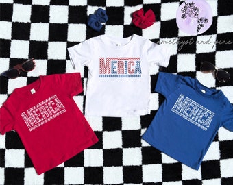 Op de afbeelding: Drie T-shirts in kindermaat met het woord "MERICA" in wit gedrukt, met rood-blauwe geruite randen. De shirts zijn rood, wit en blauw.