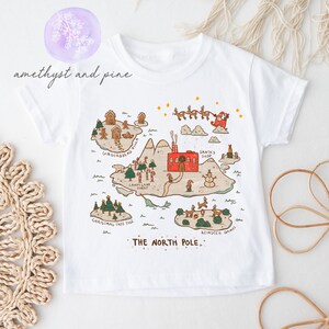May include: White t-shirt with a colorful illustration of a map to the North Pole. The map includes locations like Gingerbread Villa, Candy Cane Forest, Christmas Tree Isle, and Reindeer Games. The text "THE NORTH POLE" is at the bottom of the map.